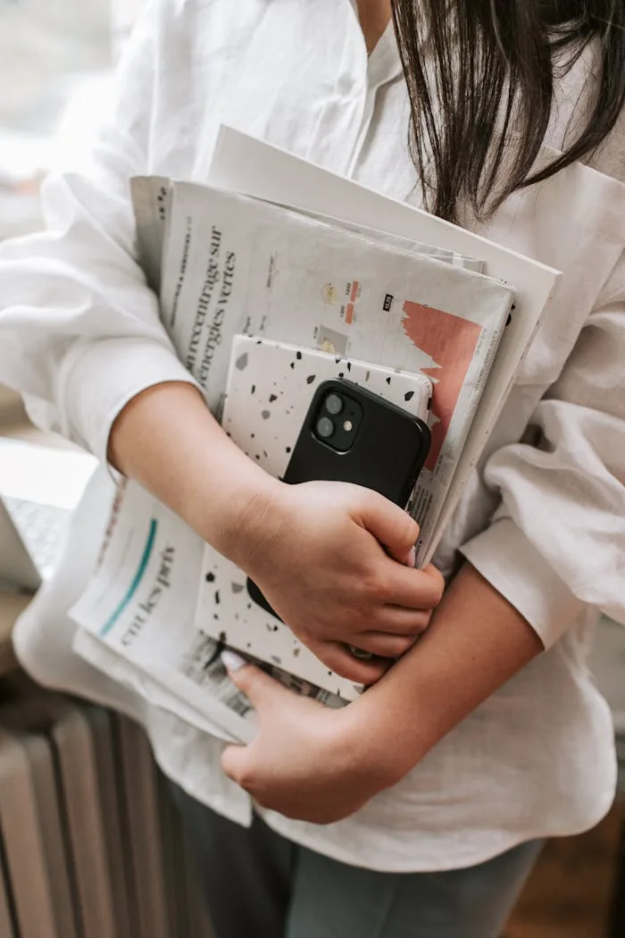 A woman in a white shirt holds newspapers and a smartphone indoors, standing by a window.